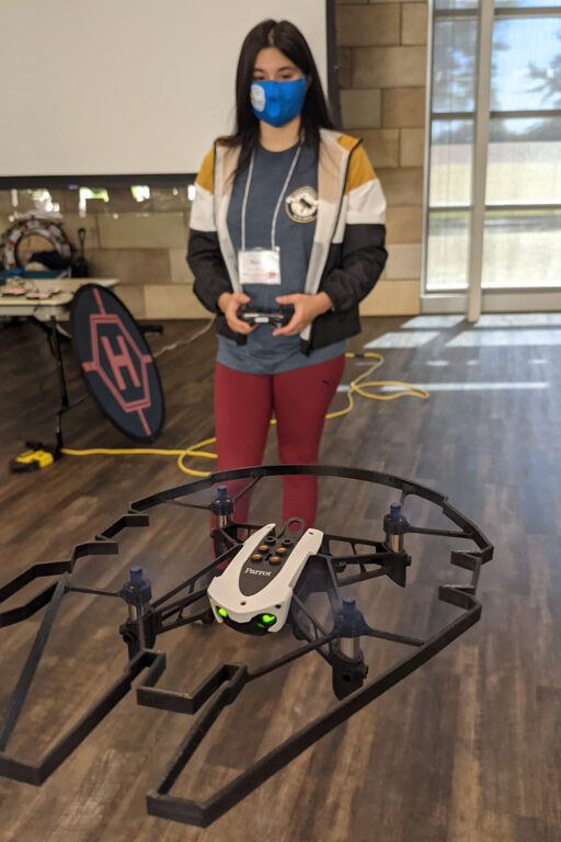 A student with long dark hair wearing a face mask pilots a mini-drone using a handset.