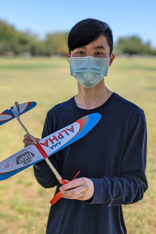 A student with short dark hair wairing a facemask holds a balsa wood glider in a field. 