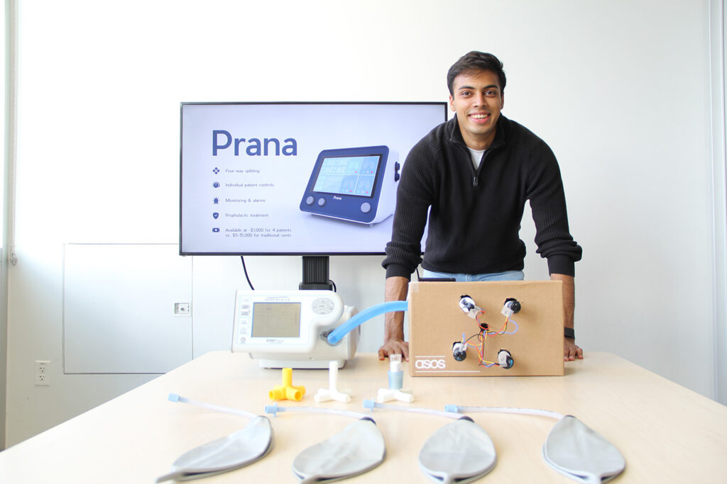 A person leans over a modified ventilator on a conference room table. In the background is a screen that reads "Prana."