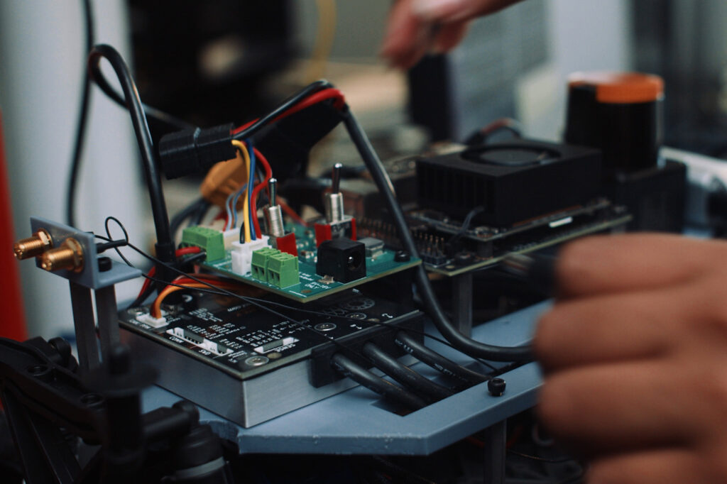 A close-up of a robotic vehicle with wires and computer chips exposed. A blurry hand reaches around one side.