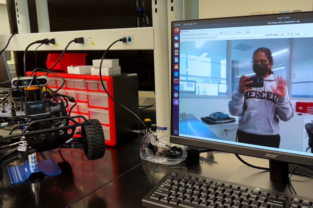A four-wheeled robot rests on a lab bench. Displayed on the computer monitor next to it is a young person wearing a UC Merced sweatshirt waving and holding a phone with the camera facing out.