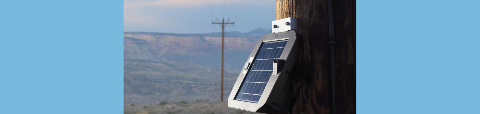 Close-up of a solar-powered sensor mounted on a powerline, with mountains in the background.