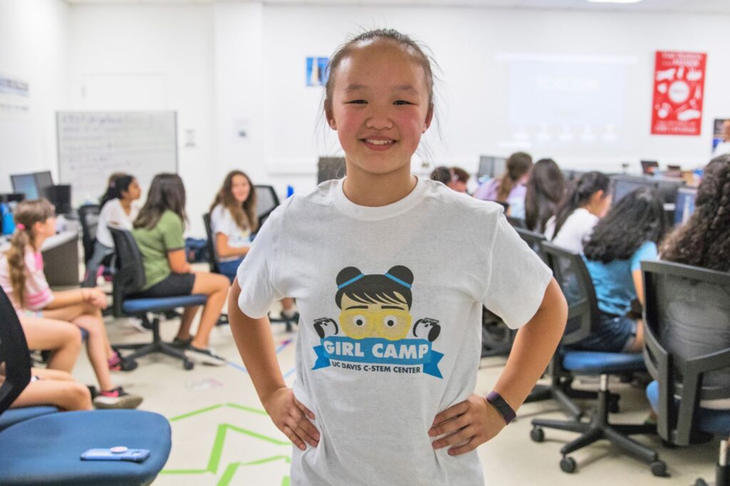A middle school-aged girl wearing a GIRL Camp t-shirt stand with her hands on her hips in a computer lab. In the background, several other girls sit at computers.