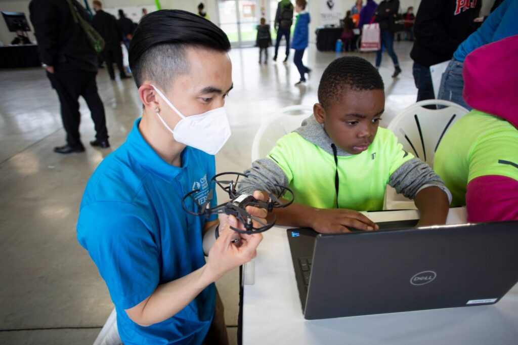A college-age person kneels, holding a small drone, next to an elementary school-aged boy who is typing on a laptop.