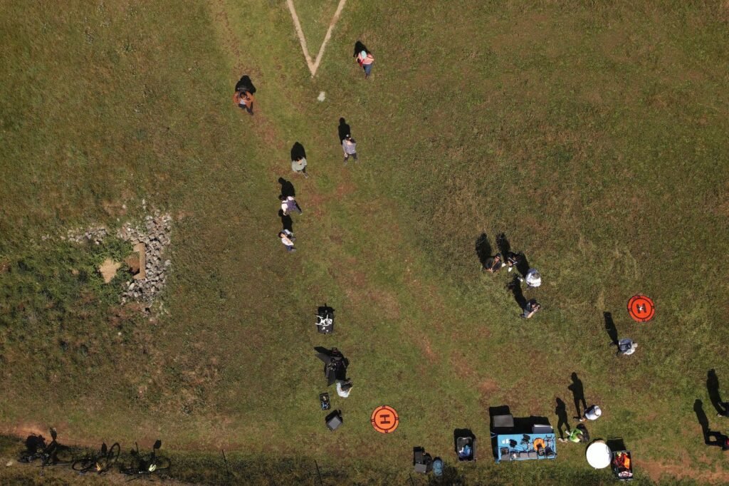 A photo from high in the air of about 14 people standing in a field, with a table holding computers in one corner and two orange targets several feet away.